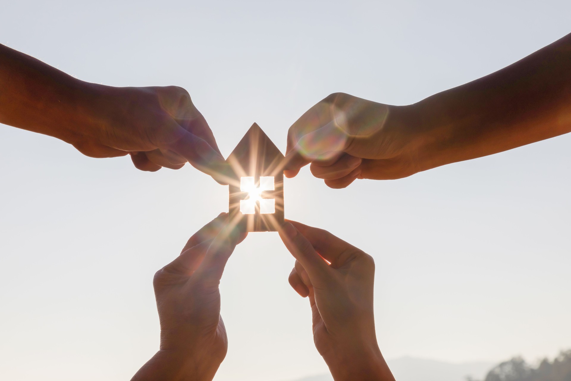 Family Hands holding wooden house toy against blue sunny sky with sun rays. solar power.